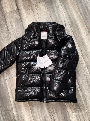 Svart glansig dunjacka från Moncler - Snygg svart dunjacka från Moncler med glansig finish och klassisk logotyp på ärmen. Jackan har huva, dragkedja framtill och praktiska fickor med dragkedja. Perfekt för kalla vinterdagar och riktigt trendig look.