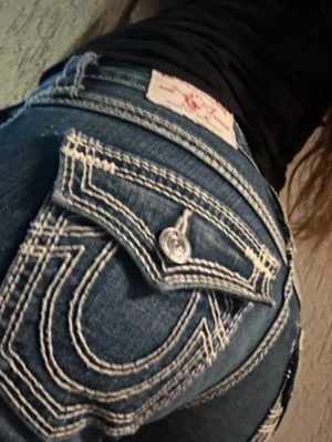 True Religion blå bootcut jeans - Säljer ett par blå jeans från True Religion med tydliga vita kontrastsömmar och stora fickor bak med dekorativa knappar. Jeansen har bootcut-modell och klassisk femficksdesign. Perfekta för dig som gillar streetwear och vill sticka ut. 