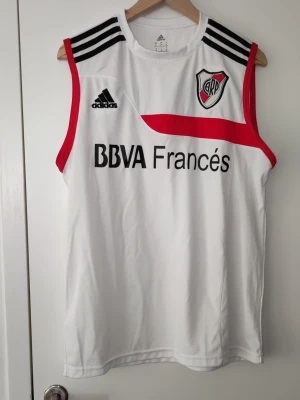 River Plate träningslinne Adidas - Säljer ett vitt träningslinne från Adidas med River Plate-logga, svarta axelränder och röda detaljer. Linnet har tryck med BBVA Francés på både fram- och baksidan. Perfekt för fotbollsträning eller gymmet.