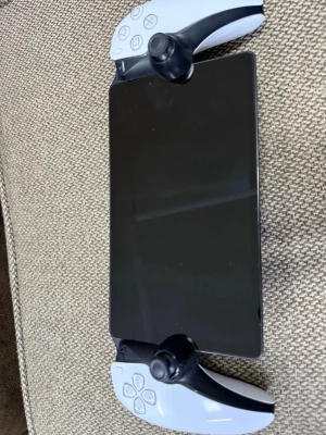PlayStation Portal handkontroll - PlayStation Portal är en handkontroll med inbyggd skärm från Sony. Den har vit och svart design, klassiska PlayStation-knappar och två analoga spakar. Perfekt för streaming av PS5-spel direkt på enheten. Materialet är plast med en matt finish och den högra styrspaken är bytt till en helt ny och fungerar perfekt.