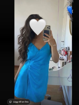 Blå kort klänning Zara  - Zara Snygg blå kortklänning med omlottdesign och smala axelband. Klänningen har rynkade detaljer i sidan och en lätt glansig yta som ger en elegant känsla. Perfekt för dig som vill sticka ut med färg! Materialet känns som polyester.