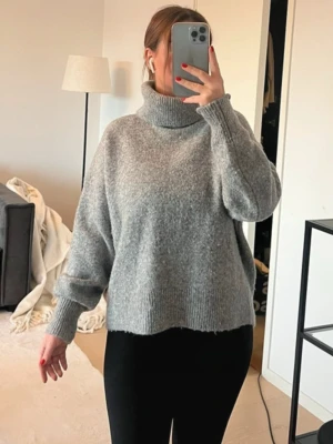 Stickad tröja  - Mysig grå stickad polotröja med lös passform och breda muddar vid ärmslut och nederkant. Tröjan har hög krage och är perfekt för kyliga dagar.