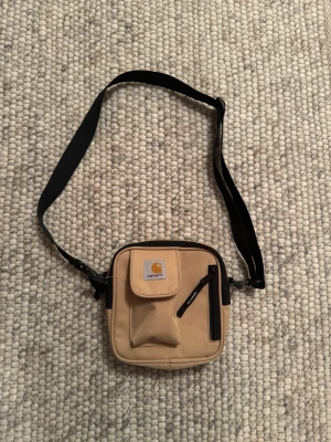  Carhartt väska - Säljer en beige axelväska från Carhartt med justerbar svart rem. Väskan har en fyrkantig form, ett litet lockfack framtill och en dragkedjeficka på framsidan. Tillverkad i slitstarkt canvasmaterial och har Carhartt-logga på locket.