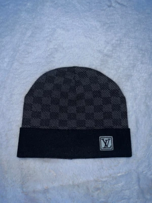 Svart mössa - Snygg svart mössa med klassiskt rutmönster och vikt kant. logga framtill på kanten ger en exklusiv touch. Perfekt accessoar för att lyfta din stil under kyliga dagar.