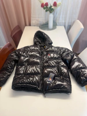 Svart dunjacka från Polo Ralph Lauren - Snygg svart dunjacka från Polo Ralph Lauren med glansig finish och huva. Jackan har dragkedja framtill, två sidofickor med dragkedja och den klassiska röda loggan broderad på bröstet. Perfekt för kalla vinterdagar.