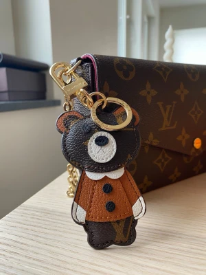  Vuitton björn nyckelring - Söt nyckelring från Vuitton i form av en björn. Tillverkad i brunt, svart och vitt läder med guldiga metalldetaljer. Perfekt att hänga på väskan eller använda till nycklar. Ikoniskt LV-mönster på björnens kropp och exklusiv känsla.