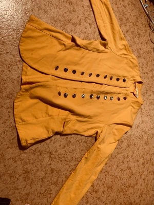 Gul jacka med dekorativa knappar - Snygg gul jacka i bomull med unika silverfärgade knappar framtill. Jackan har lång ärm och fickor på sidorna. Perfekt för dig som vill sticka ut med färg och detaljer.