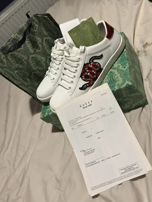 Gucci vita sneakers med orm - Säljer ett par vita sneakers från Gucci med den ikoniska röda och svarta ormen broderad på sidan. Skorna har snörning, rund tå och platt sula. Kommer med originalkartong och påse. Perfekta för dig som vill sticka ut med lyxig streetstyle.strl 41/42 använda endast en gång endast box följer med