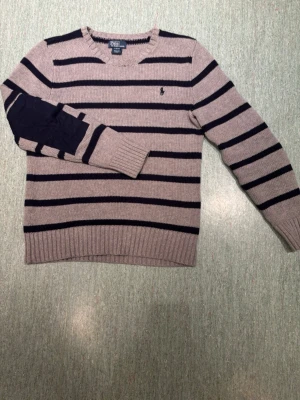 Randig stickad tröja från Polo Ralph Lauren - Snygg stickad tröja från Polo Ralph Lauren. Grå med mörkblå horisontella ränder. Tröjan har rund halsringning, långa ärmar och mörkblå patch på armbågarna. Klassisk logga broderad på bröstet. Perfekt för en stilren look.