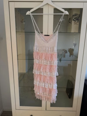 Rosa och vit spetsklänning med volanger - Supersöt kort klänning med tunna axelband i rosa och vitt. Klänningen har flera lager av spetsvolanger och v-ringad urringning. Perfekt för sommarens alla tillfällen och ger en romantisk vibe.