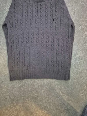 Grå kabelstickad tröja Polo Ralph Lauren - Kabelstickad grå tröja från Polo Ralph Lauren med rund halsringning och diskret broderad logga på bröstet. Tröjan har ribbstickade muddar vid ärmslut och nederkant. Perfekt för lager-på-lager och klassisk stil.