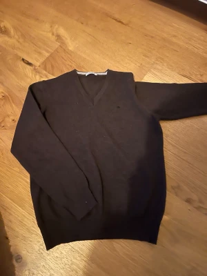 Mörkbrun stickad tröja från J.Lindeberg - Snygg mörkbrun stickad tröja från J.Lindeberg med V-ringning och  broderad logga på bröstet. Tröjan har långa ärmar och ribbade muddar vid ärmslut och nederkant. Perfekt för lager på lager och stilrena outfits. storlek 158-164 och lite större