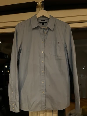 Ljusblå skjorta från Tommy Hilfiger - Klassisk ljusblå skjorta från Tommy Hilfiger med bröstficka och liten logga på fickan. Skjortan har knappar framtill och lång ärm. Tillverkad i bomull och har en normal passform. Perfekt för en clean och stilren look.