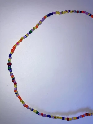 Färgglatt pärlhalsband - Ett handgjort halsband med små, färgglada plastpärlor i olika nyanser som rött, blått, gult, grönt och lila. Halsbandet har en silverfärgad kedja och karbinlås för enkel justering. Perfekt för dig som vill addera en lekfull touch till din stil.