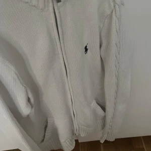 Vit stickad kofta från Ralph Lauren - Säljer en stilren vit stickad kofta från Ralph Lauren med dragkedja och ribbade detaljer. Koftan har ett diskret broderat logotyp på bröstet och flätade detaljer längs ärmarna. Perfekt för kyliga dagar!