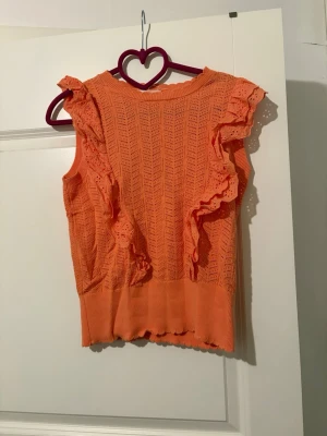 Orange ärmlös topp med volang - Snygg orange ärmlös topp med virkade detaljer och volanger längs axlarna. Toppen har ett mönstrat stickat tyg och bred ribbad kant nedtill. Perfekt för dig som vill ha en färgglad och unik look.
