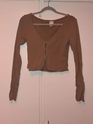 Beige croppad ribbad topp med knappar - Trendig beige croppad topp med lång ärm och v-ringning. Toppen är ribbad och har knappar framtill för en snygg detalj. Perfekt att styla med höga jeans eller kjol för en chill look.