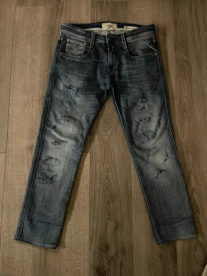 Replay slitna blå jeans herr (uppsydda) - Säljer ett par blå jeans från Replay med slitningar, jeansen är kapade hos jeansbolaget från 90cm till 83 cm passar en som är runt 155-160, femficksmodell med raka ben och snygg tvättad look. Perfekta för en avslappnad och trendig stil hör av er för frågor.