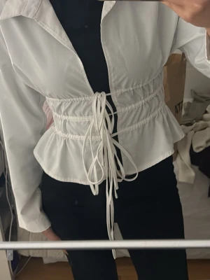 Vit croppad blus med snörning - Snygg vit croppad blus med långa ärmar och markerad midja med snörning framtill. Blusen har klassisk krage och volangkant nedtill för en trendig look. Perfekt att styla över en topp för en cool outfit.