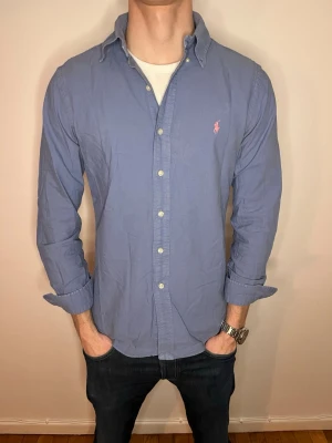 Blå Ralph Lauren skjorta  - Klassisk blå skjorta från Ralph Lauren med button-down krage och den ikoniska rosa logotypen broderad på bröstet. Skjortan har långa ärmar, knappar framtill och är tillverkad i mjuk bomull. Perfekt för en clean och stilren look. Skicket 10/10 och pris 275 men kan gå ner vid snabb och smidig affär💙📦Storlek S men passar M Hör av er vid frågor, vi postar så snabbt som möjligt🥰
