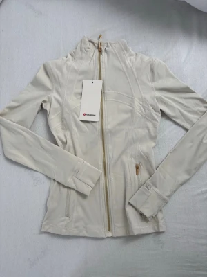 Vit defyne jacket från lululemon - Snygg oanvänd dfyne jacka med guld dragkedja från lululemon helt ny med prislapp
