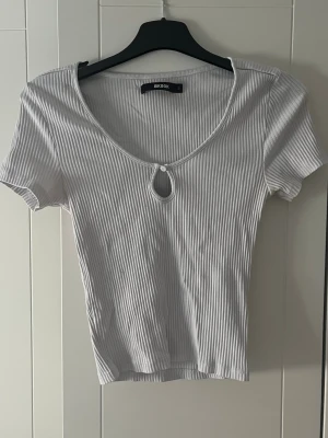 Vit ribbad topp från Bik Bok - Kortärmad, croppad topp från Bik Bok i vit ribbad bomull. Toppen har en v-ringad halsringning med en liten cut-out detalj framtill. Perfekt för en trendig och avslappnad stil.