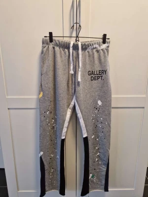Grå mjukisbyxor Gallery Dept. - Grå mjukisbyxor från Gallery Dept. med svart och vit kontrast längs benen, elastisk midja med vit dragsko och tryckt logga på vänster lår. Färgstänk och detaljer ger en unik, kreativ vibe. Perfekta för en avslappnad och trendig stil.