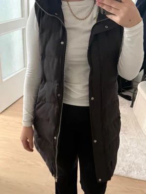 Svart lång dunväst med huva - Snygg svart lång dunväst med dragkedja och tryckknappar framtill. Västen har en stor huva och quiltad design som ger en modern look. Perfekt att slänga över en hoodie eller tröja för extra värme. Passar perfekt nu till våren. Flitigt använd men ff i relativt bra skick!!