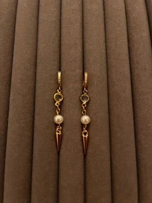 Guldörhängen med pärlor och spets - Snygga örhängen i guldton med små vita pärlor och spetsiga hängen. Örhängena har en rund detalj upptill och stängs med klicklås. Perfekta för dig som gillar en mix av klassiskt och edgy stil.