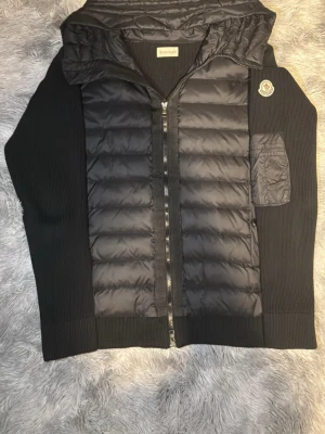 Moncler Cardigan - Snygg svart dunväst från Moncler med quiltad front, stickade partier på sidor och huva. Västen har dragkedja, fickor med knapp och en Moncler-logga på bröstet. Perfekt för lager-på-lager och streetstyle. Om du känner dig intresserad så är det bara ställe era frågor till mig pris går även att diskutera 