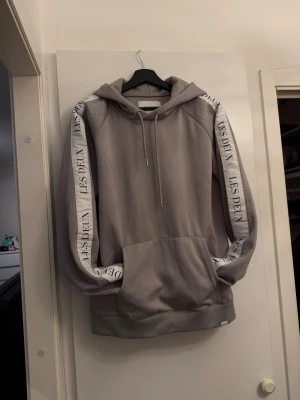 Beige hoodie från Les Deux med tryck - Snygg beige hoodie från Les Deux med huva och dragsko. Har vita band längs ärmarna med svart Les Deux-tryck. Stor känguruficka framtill och ribbade muddar. Perfekt för en chill och stilren look. I mycket bra skick, inga skador.