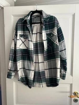 Grönrutiga overshirt från Bik Bok - Overshirt i grönt, vitt och svart rutmönster från Bik Bok. Modellen har två bröstfickor med knappar, klassisk krage och långa ärmar. Perfekt att slänga över en t-shirt för en avslappnad look. Materialet är mjukt och känns som flanell.