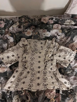 Blommig blus med volanger - Söt beige blus med bruna blommönster och volangdetaljer längs framsidan. Blusen har v-ringning och trekvartsärm, vilket ger en avslappnad men ändå trendig look. Perfekt att styla med jeans eller kjol och mycket bra till sommarkvällar ❤️