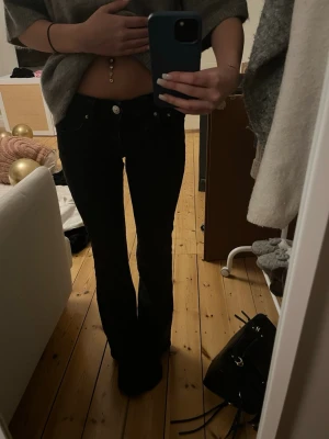 Svarta bootcut jeans med låg midja - Säljer ett par svarta bootcut jeans med låg midja och klassisk femficksdesign. Jeansen har silverfärgade knappar och smal passform upptill som går ut i ett lätt utsvängda ben. Perfekta för en trendig och avslappnad look.