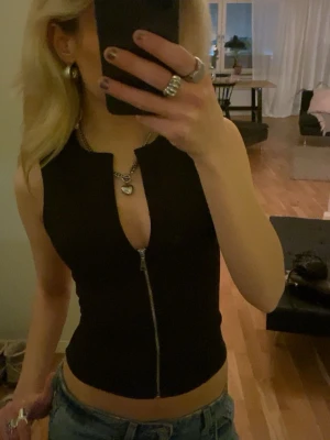 Svart ärmlös topp med dragkedja - Snygg svart ärmlös topp med hel dragkedja framtill och en liten v-ringning vid halsen. Toppen är croppad och har en tight passform, perfekt att styla med jeans eller kjol. Enkel och stilren design utan synligt märke.
