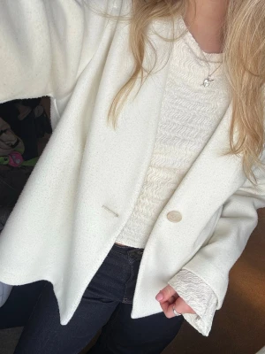 Vit oversized kappa med knapp - Stilren vit kappa i oversized modell med en enkel knappstängning framtill. Kappan har rak passform, långa är ärmar och sidofickor. 