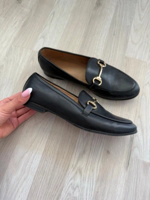 Svarta loafers med gulddetalj - Stilrena svarta loafers i slätt läder med klassisk guldfärgad metallspänne ifrån hm i storlek 37💕