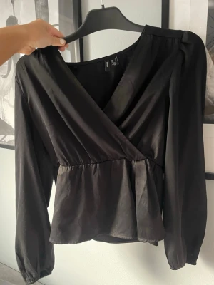 Svartblus  - Stilren svart omlottblus från Veromoda med v-ringning och markerad midja. Blusen har långa ärmar med lätt puff vid axlarna och resår vid ärmsluten. Perfekt att matcha med både jeans och kjol för en trendig look.💕💕💖Säljer då den inte längre används. 