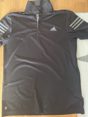 Svart pikétröja från Adidas (golf) - Svart pikétröja från Adidas med klassiska tre ränder på ärmarna och logga på bröstet. Tr drawn har knappar vid kragen och är gjord i ett syntetmaterial som passar perfekt för träning eller sportiga aktiviteter. (Golf)