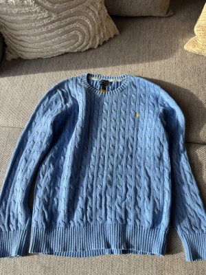Ljusblå kabelstickad tröja Polo Ralph Lauren - Snygg ljusblå kabelstickad tröja från Polo Ralph Lauren med rund halsringning och gul broderad logga på bröstet. Tröjan har ribbade muddar vid ärmslut och nederkant och är tillverkad i mjukt material. Perfekt för en chill och stilren look.