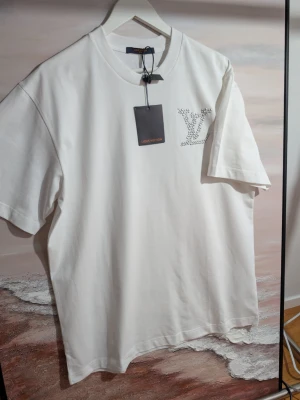 Iouis Vuitton T-shirt  - Vit t-shirt med pärldetaljer och orange läderpatch Exklusiv vit t-shirt i mjuk bomullstrikå med unika detaljer. Framsidan pryds av en mindre logotyp på bröstet skapad av vita pärlor, medan baksidan har en stor och iögonfallande logotyp samt text i samma pärlarbete. En stilren orange läderpatch vid nacken ger en extra lyxig touch. Färg: Vit med pärlvita detaljer. Design: Rundhalsad, korta ärmar och avslappnad passform. Detaljer: Broderade pärlor med 3D-känsla och signaturpatch i läder på ryggen.
