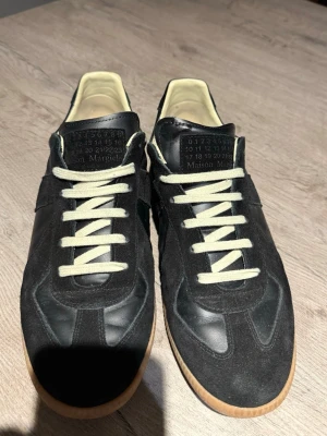 Svarta Maison Margiela gats - Maison Margiela sneakers i svart läder och mocka med vita snören och beige innersula. Klassisk låg modell med gummisula och ikonisk logotyp på plösen. Snygg kontrast mellan material och färger, perfekt för en stilren streetwear-look. Använda 5 gånger typ 