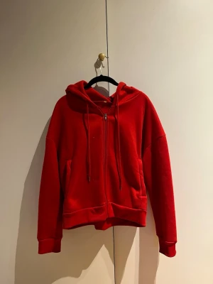 Röd hoodie med dragkedja -  sopig röd hoodie med dragkedja framtill och huva med snören. Hoodien har två fickor på sidorna och ribbade muddar vid ärmslut och nederkant. Perfekt för en chill och avslappnad stil.