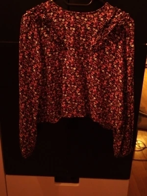 Blommig långärmad blus med volang - Blus med svart botten och små blommor i orange, rött och vitt. Långärmad modell med volangdetalj vid axlarna och lös passform. Perfekt för dig som gillar färgglada mönster och en romantisk stil. Materialet känns mjukt och följsamt. Det är en matchande tjol till.