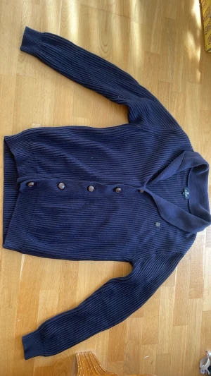 Ribbad cardigan  - Snygg mörkblå ribbstickad kofta från polo med bruna knappar framtill och bruna mockaimitation-lappar på armbågarna. Långärmad modell med bred krage och ribbade muddar. Perfekt för dig som gillar klassisk trailingstil. Riktigt skön nu inför våren, strl L