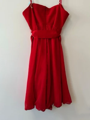 Röd kort klänning med knytband - Snygg röd klänning med tunna axelband och ett brett knytband i midjan. Klänningen har en luftig kjol med lätt volang och är tillverkad i ett tunt, flygigt material som ger en elegant känsla. Perfekt för dig som vill sticka ut med färg. Storlek L men sitter som S. 