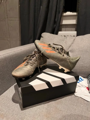 Adidas grå-orange fotbollsskor  - Säljer ett par grå fotbollsskor från Adidas med klassiska orangea ränder på sidorna och snörning. Skorna har låg profil och är tillverkade i syntetmaterial för bra grepp och kontroll på planen. Perfekta för dig som vill sticka ut på matchen!