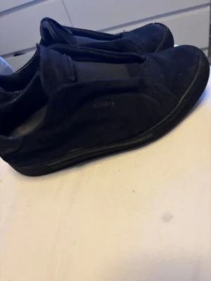 Svarta sneakers från Arigatos söker byten  - Snygga svarta sneakers från Arigatos i mocka med minimalistisk design. Skorna har en låg silhuett och diskret logga på sidan. Perfekta för dig som gillar stilrena och klassiska sneakers med en modern touch.