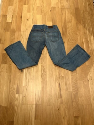 Blå bootcut jeans från Lee - Säljer ett par klassiska blå bootcut jeans från Lee med fem fickor och normal midja. Jeansen har en snygg tvättad look och är tillverkade i mjukt denimtyg. Perfekta för dig som gillar en avslappnad men trendig stil. Jag hjälper min tjej att sälja hennes kläder!