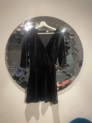 Svart sammetsklänning med v-ringning - Kort svart sammetsklänning med djup v-ringning och trekvartsärm. Klänningen har omlott-design either framtill och markerad midja för en snygg siluett. Perfekt för dig som vill ha en elegant och trendig look.